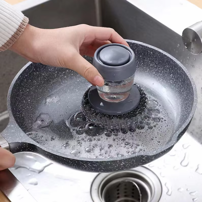 Person using a soap dispenser steel brush to clean a frying pan in a sink.